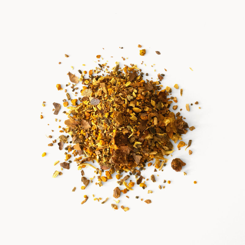 Against a white background lies a small pile of loose herbal tea leaves and spices from Rishi Tea & Botanicals' Pumpkin Spice blend, featuring an array of dried herbs and spices in shades of brown, yellow, and green that promise a chai-like infusion with hints of seasonal botanicals.