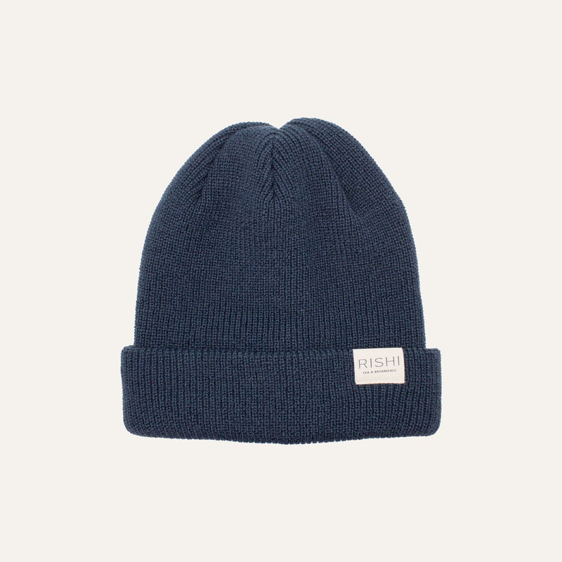 The dark blue Rishi Ribbed Beanie by Rishi Tea & Botanicals features a folded brim and a small rectangular white label with text. Crafted from recycled acrylic, it is Fair Trade Certified for ethical craftsmanship.