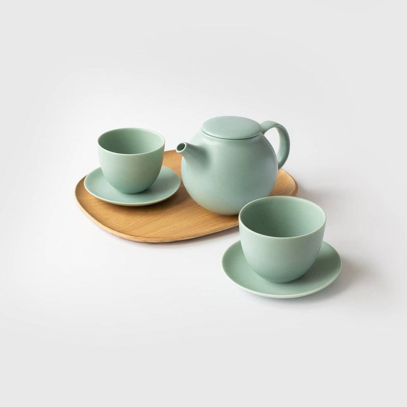 A light green Rishi Tea & Botanicals Pebble Porcelain Tea Set, with a teapot, matching teacups, and saucers, is elegantly arranged on a non-slip wood serving tray against a plain background.