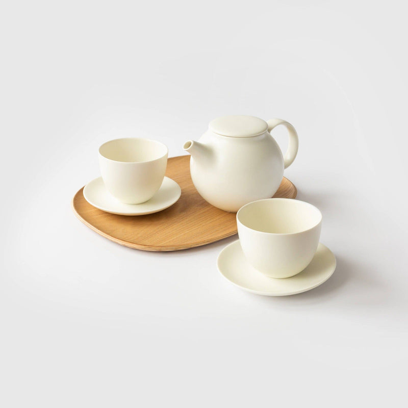 A Rishi Tea & Botanicals tea set adorns the plain background, showcasing a white teapot with a handle on a wooden serving tray, accompanied by two immaculate white teacups and saucers for an inviting tea experience.